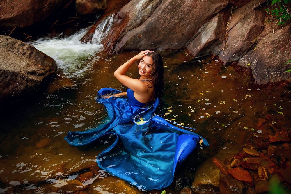Artistic photoshoot in a forest stream featuring a flowing blue dress and natural rock formations.