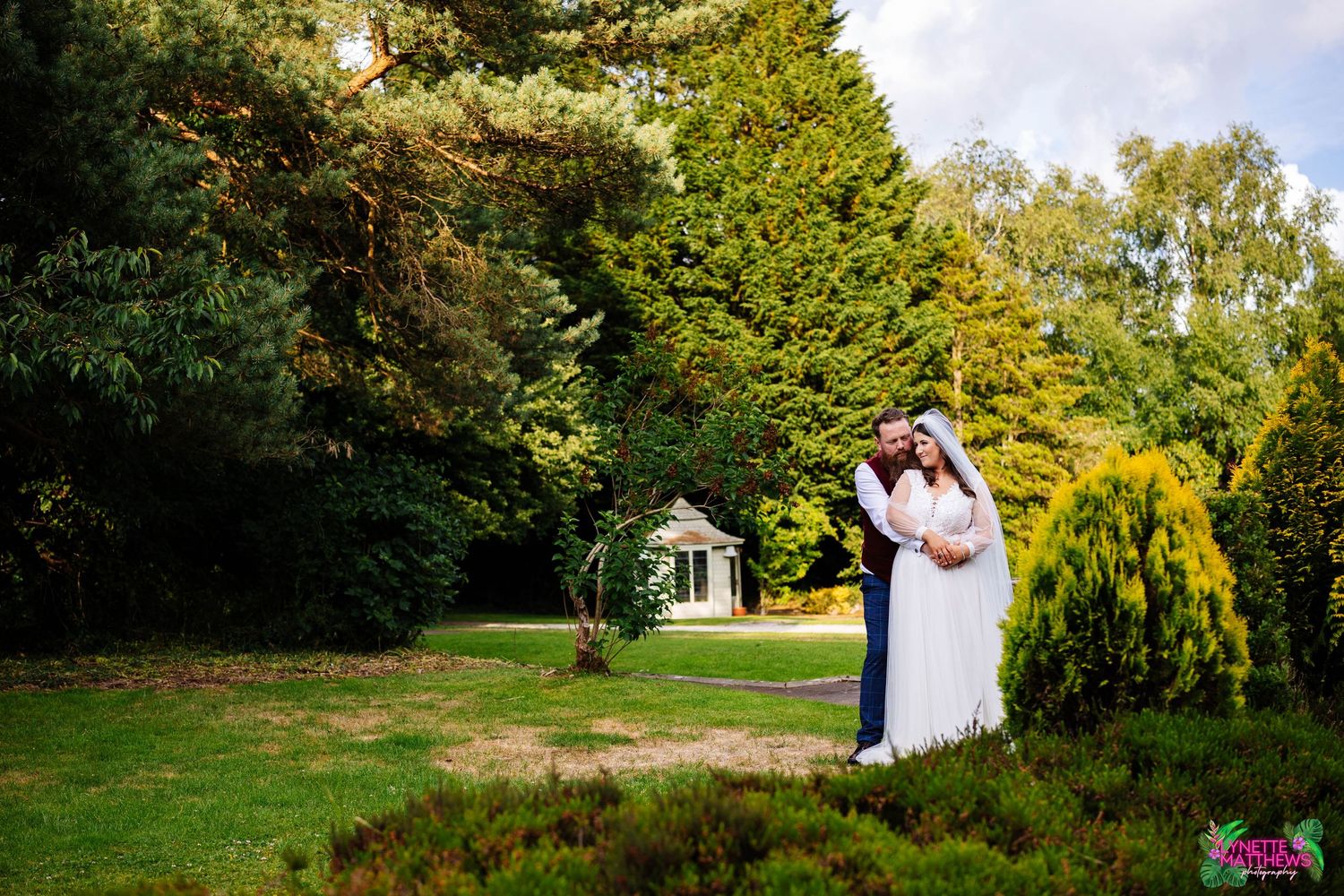 A bride and groom embraces on a lush green lawn with trees & a white gazebo in the background at Bartle Hall by lynette matthews photography