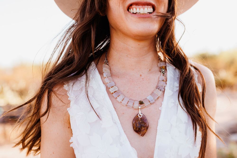 Woman in white sleeveless top wearing layered bohemian necklaces with crystal pendant smiles in sunlit field.