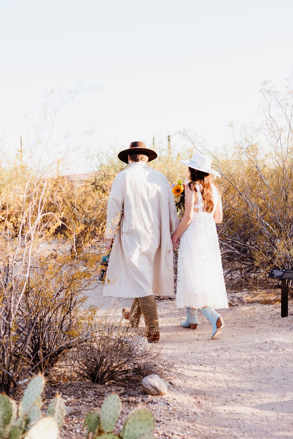 A couple in all white attire walking on a desert trail surrounded by cacti and desert brush at sunset.