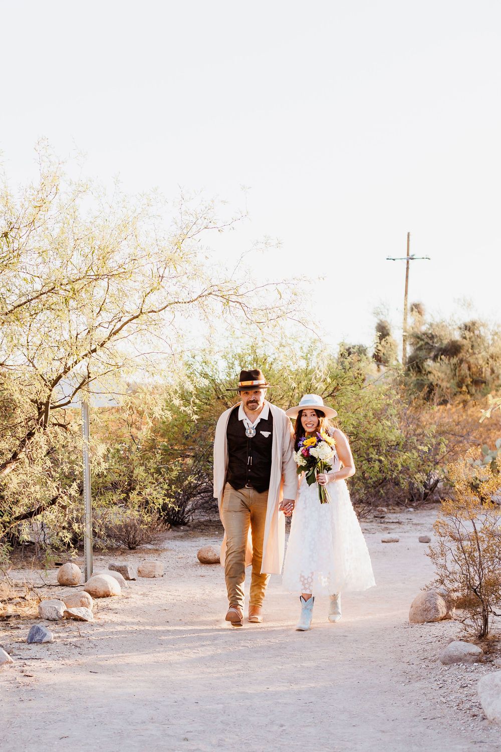 A couple walks hand in hand down a desert path surrounded by shrubs and rocks on a sunny afternoon.