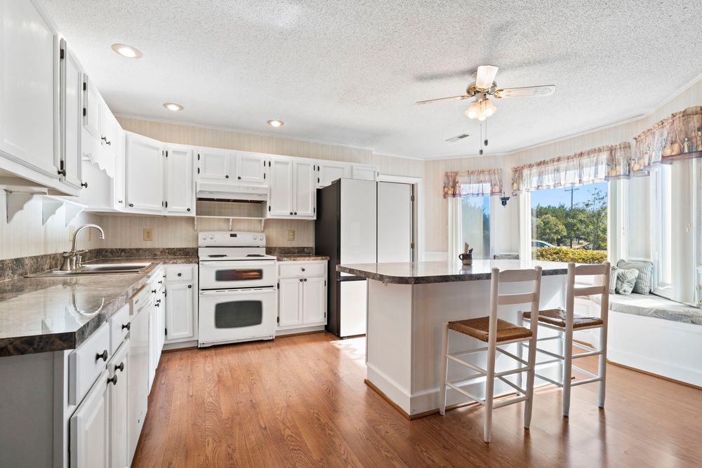 Classic white kitchen with wood floors, center island with seating, granite counters, and sunny bay window.