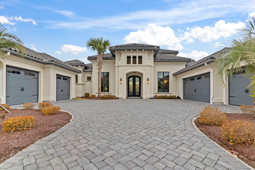 Elegant Mediterranean-style home with a paver driveway, triple garage, arched entry, and lush tropical landscaping.