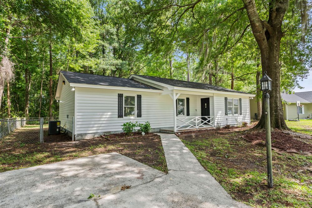 Charming white ranch-style home with a covered porch, concrete walkway, and mature shade trees in a wooded setting.