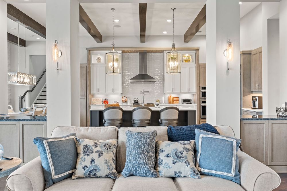 Bright open-concept living room with beamed ceilings, neutral sofa with blue pillows, and gourmet kitchen beyond.