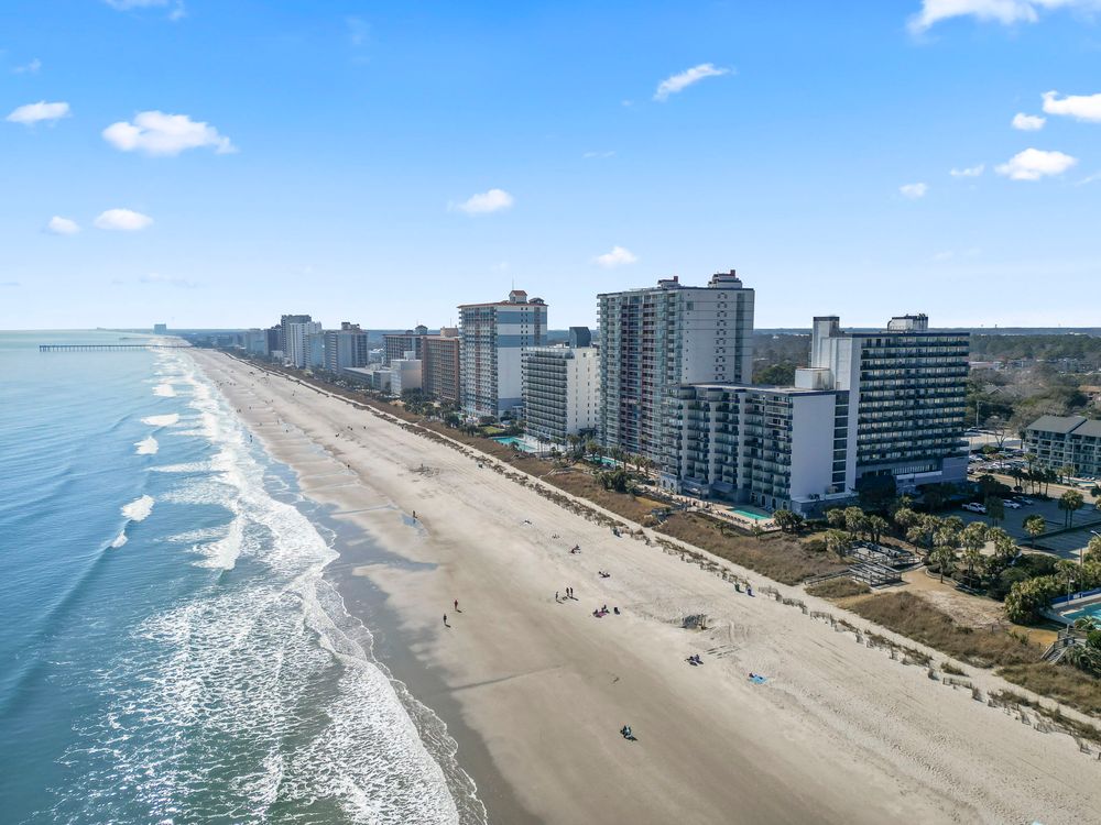 Aerial view of a wide sandy beach with ocean waves and coastal high-rise hotels stretching into distance.