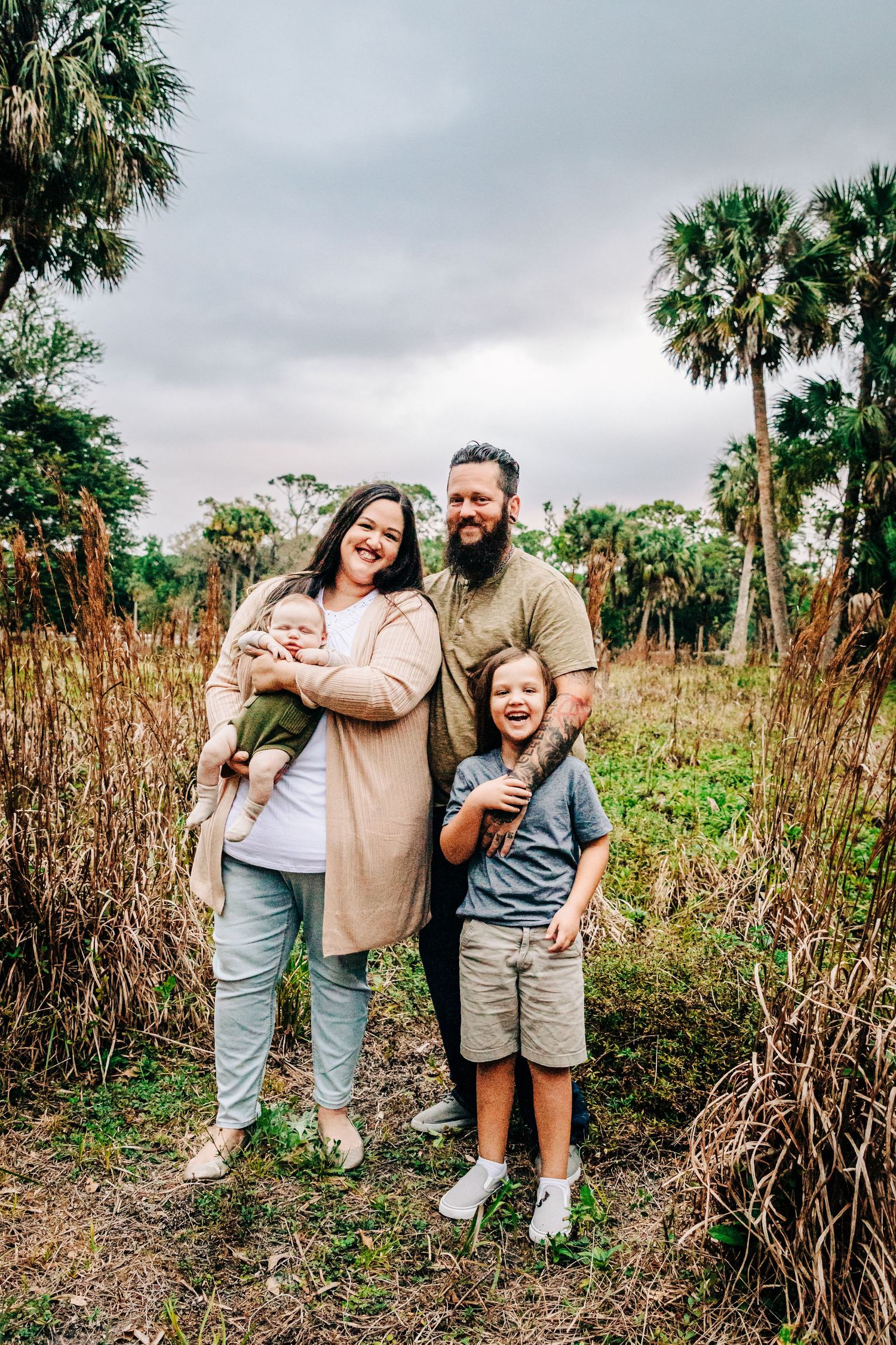 A family stands together outdoors surrounded by tall palm trees and grassy vegetation on an overcast day at riverbend park in Jupiter fl.