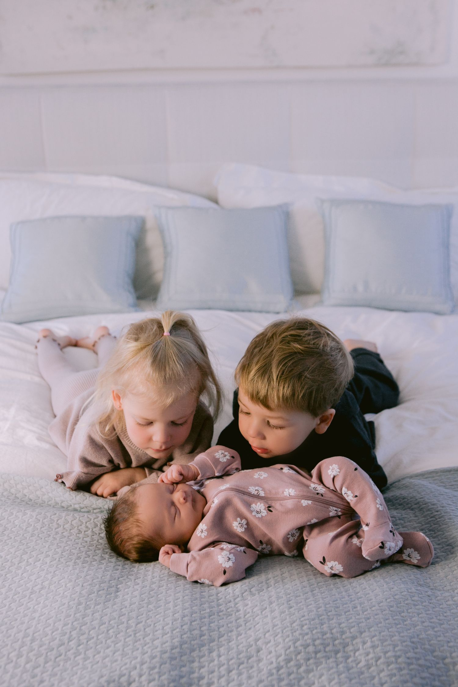 Siblings cuddling with newborn baby on white bedding in natural light.
