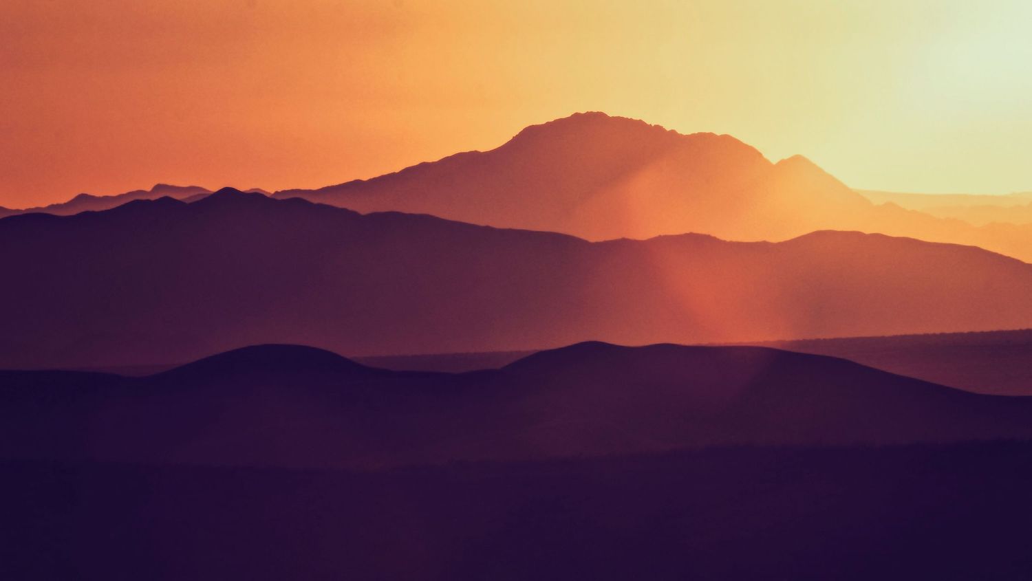 Warm orange sunset light illuminates layered desert mountain silhouettes with golden rays.