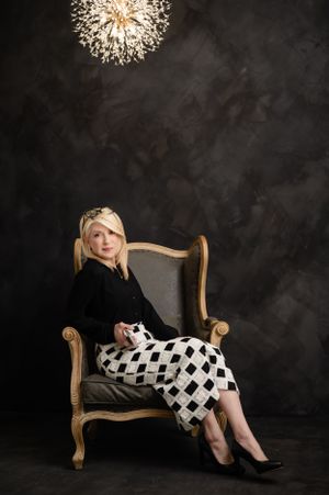 Professional portrait with elegant black and white checkered outfit on vintage chair under modern light fixture.