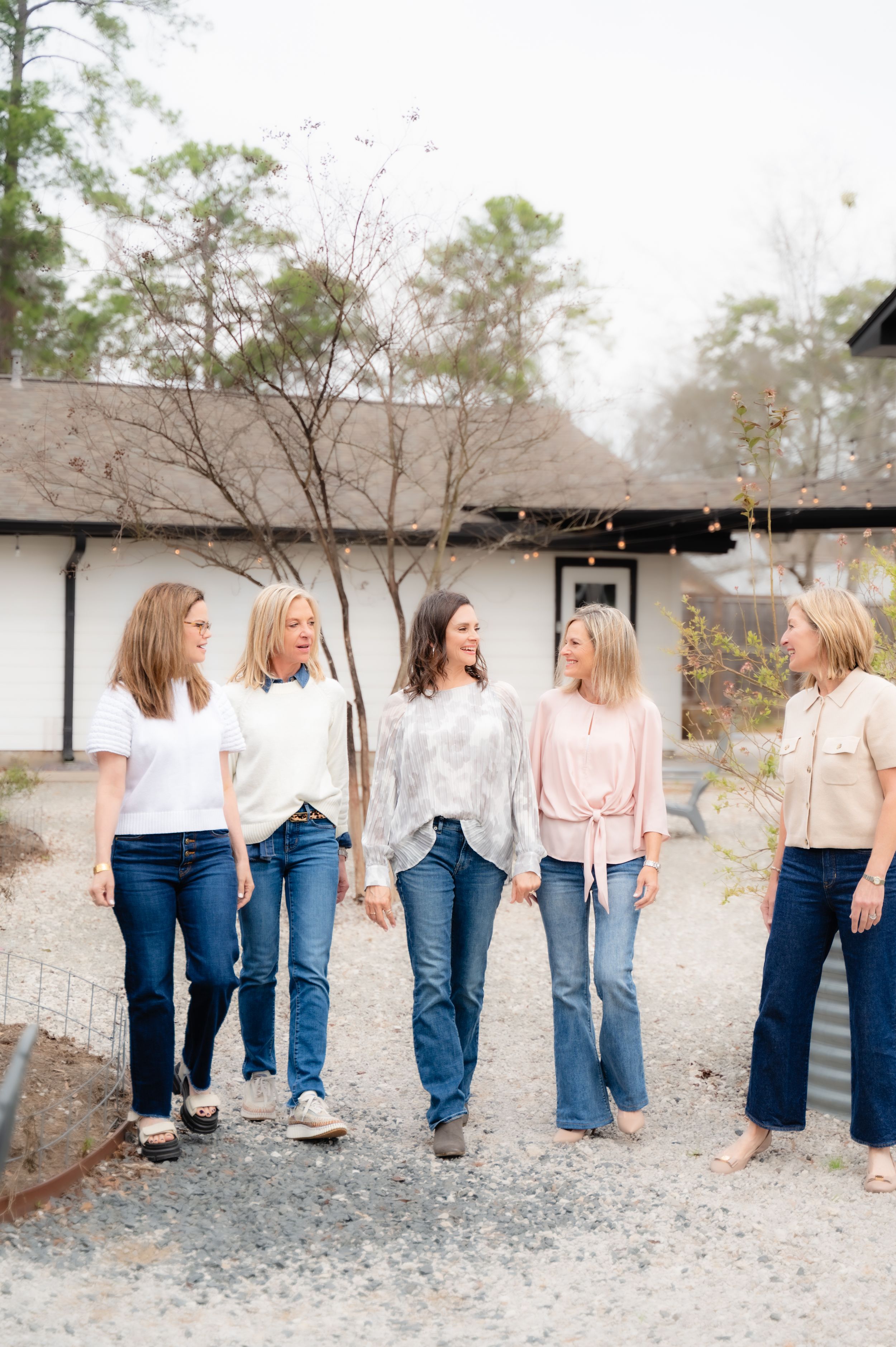 Five women in casual denim and neutral tops walk together on a gravel path outside a rustic building.