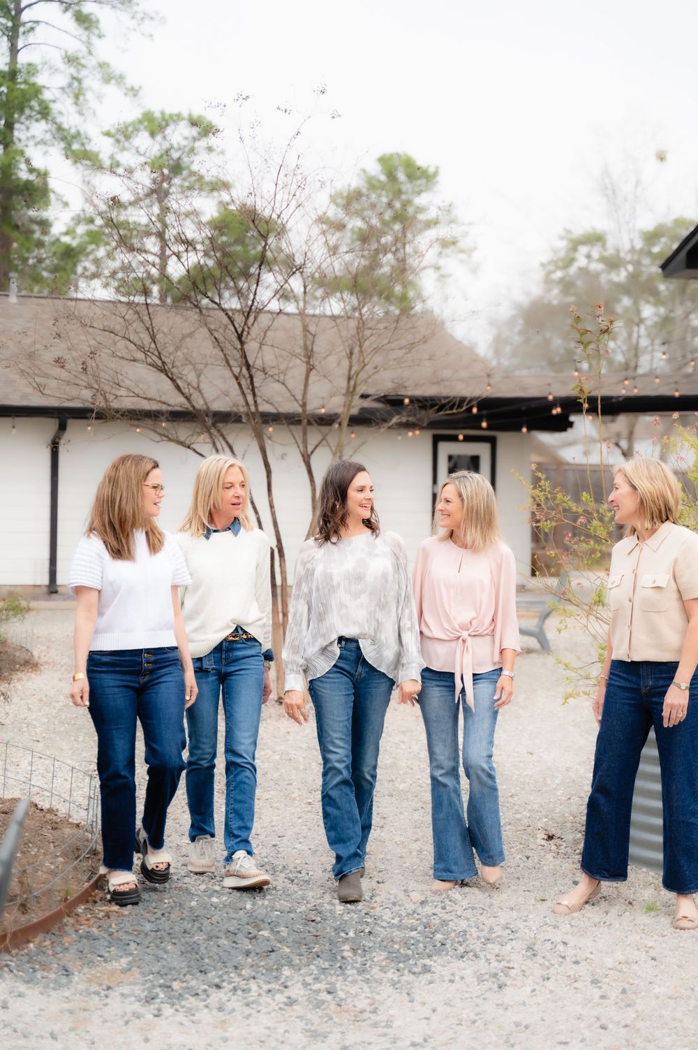 Five women in casual denim and neutral tops walk together on a gravel path outside a rustic building.