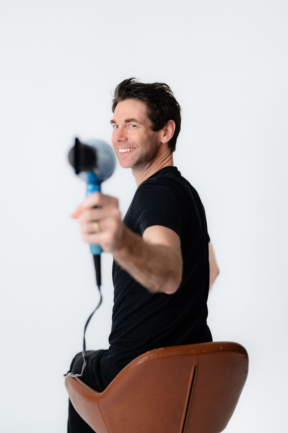 Person in black t-shirt sits on brown leather chair holding a blow dryer while smiling.