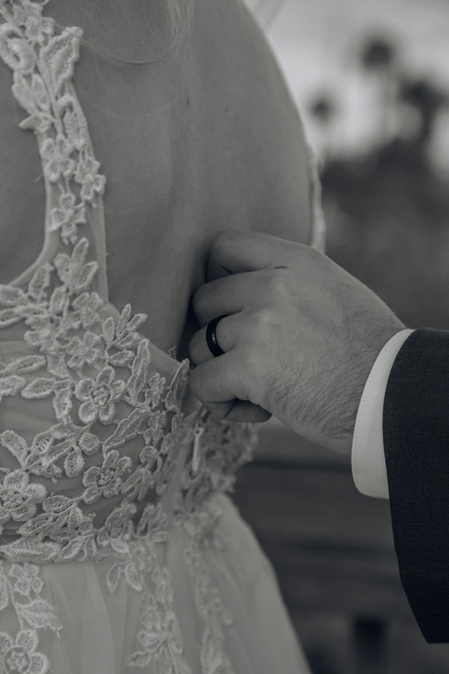 Black and white close-up of groom fastening lace wedding dress buttons on bride.