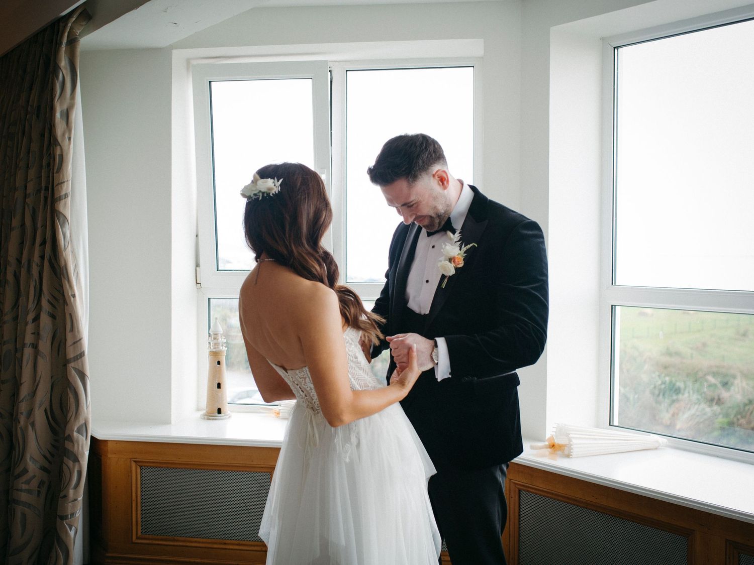 A couple in wedding attire stands in front of a bright window taking photos during a romantic moment.