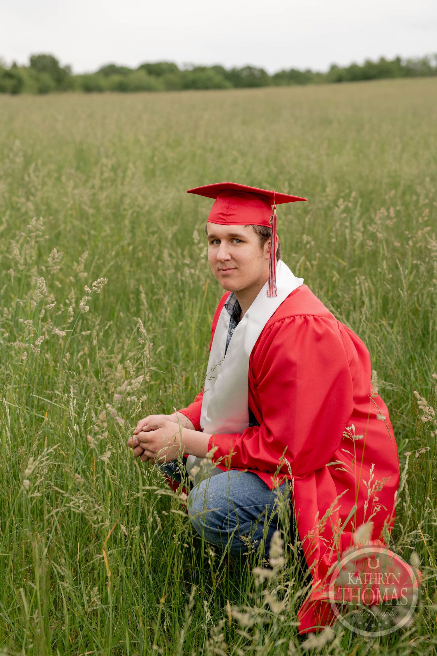 Graduate in bright red cap and gown kneels in a tall grass field during outdoor photo session.