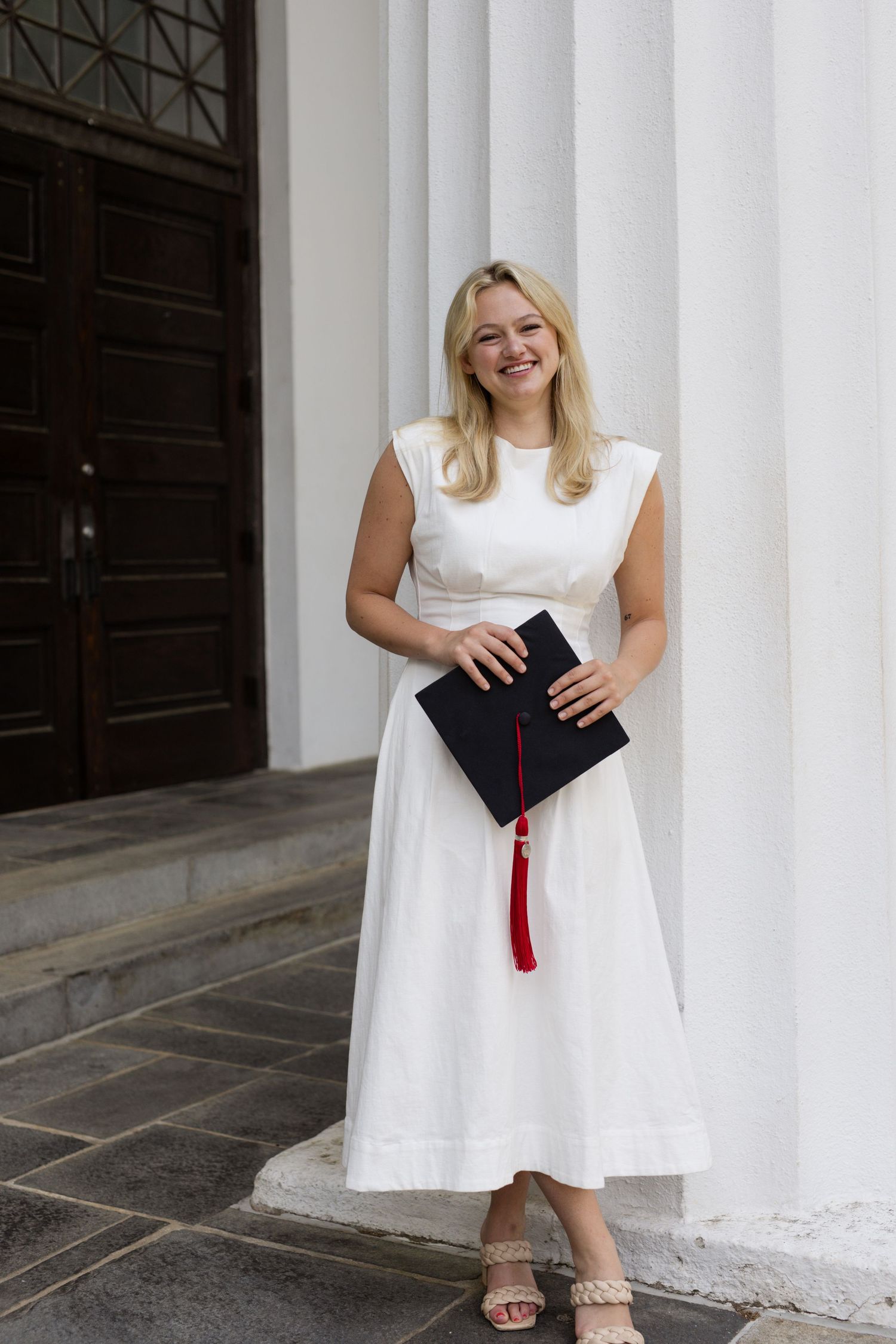 Graduate in white dress poses with diploma and tassel against white columns.