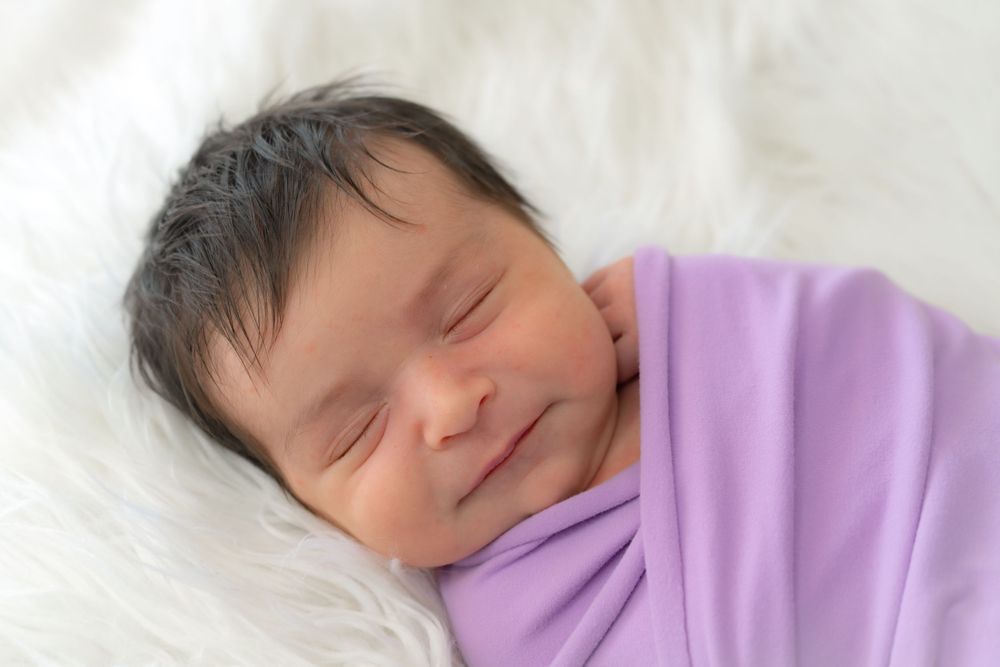 Sleeping newborn wrapped in soft purple blanket rests peacefully on white fur background.