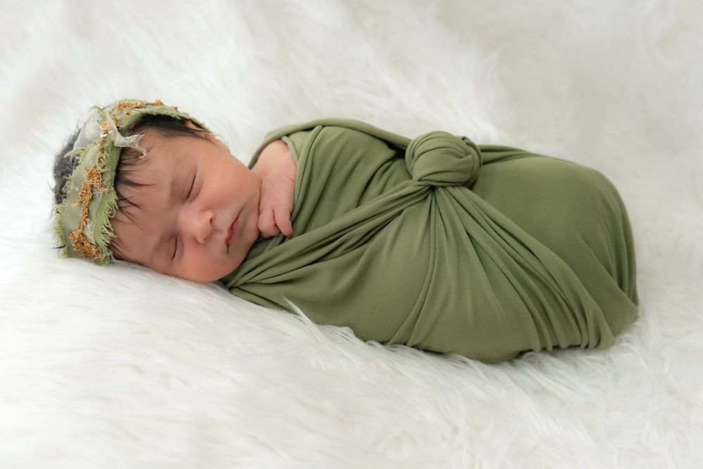 Sleeping newborn baby wrapped snugly in olive green swaddle blanket lying on white fluffy backdrop with delicate headband.
