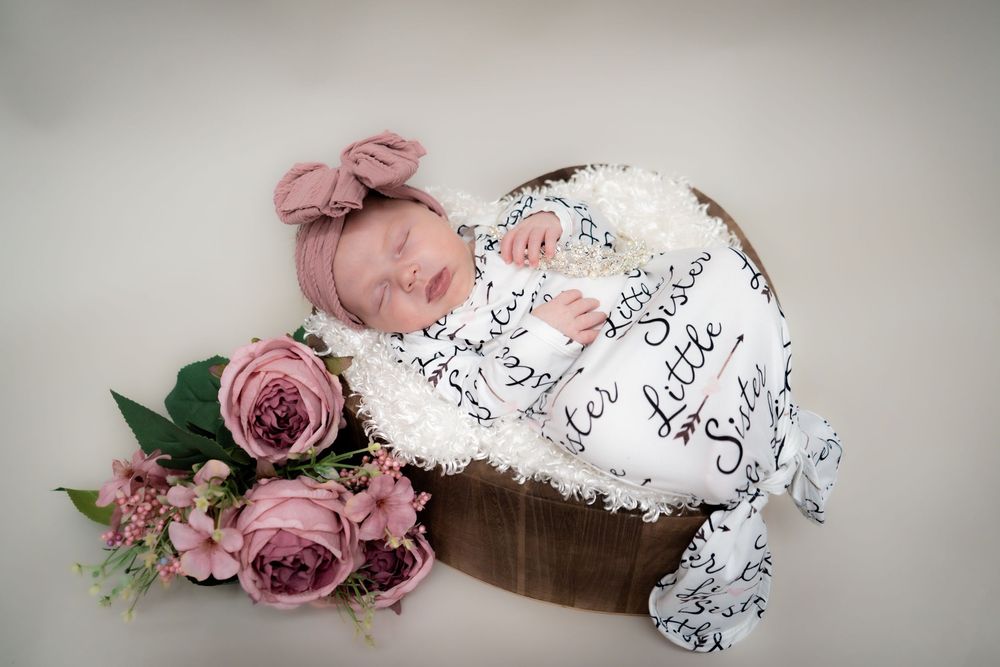 Sleeping newborn wrapped in musical note swaddle blanket lies in wooden bowl beside dusty pink roses.