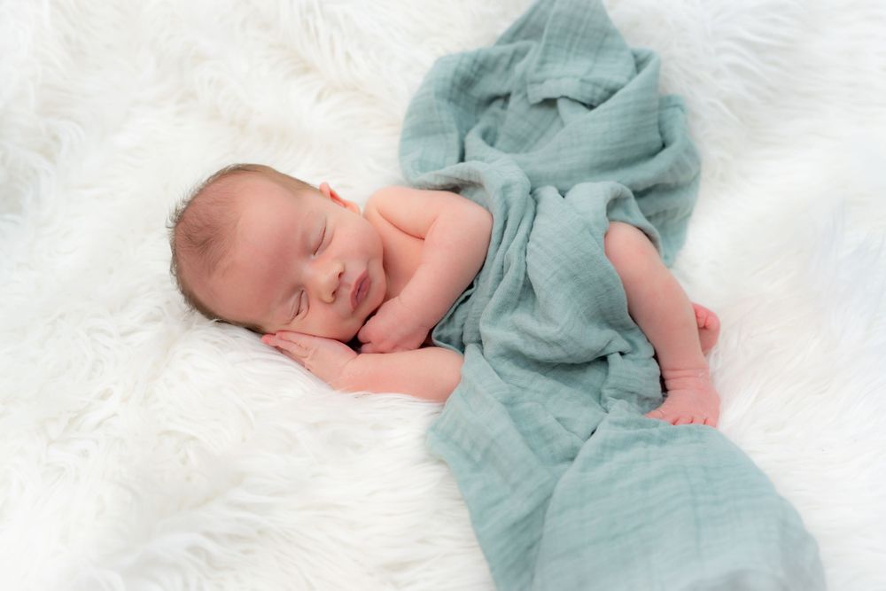 Sleeping newborn wrapped in soft mint green fabric on white fur backdrop.