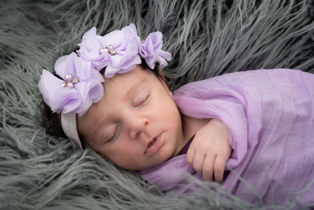 A sleeping infant wearing purple pajamas rests peacefully on a soft gray fur blanket.