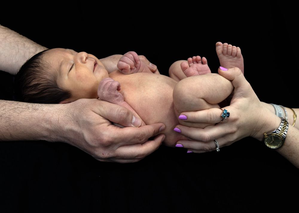 A newborn baby is cradled and supported by adult hands against a black background.