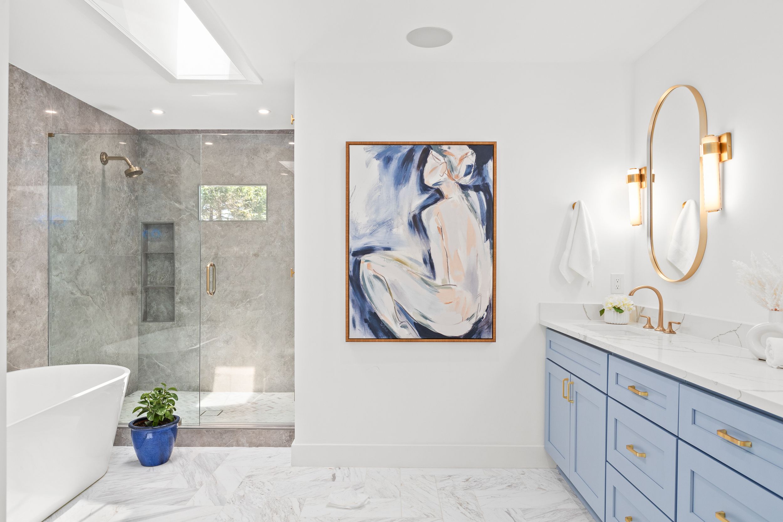 Modern bathroom with marble floors, blue vanity cabinet, gold fixtures, and abstract blue artwork on white walls. Real estate photography.