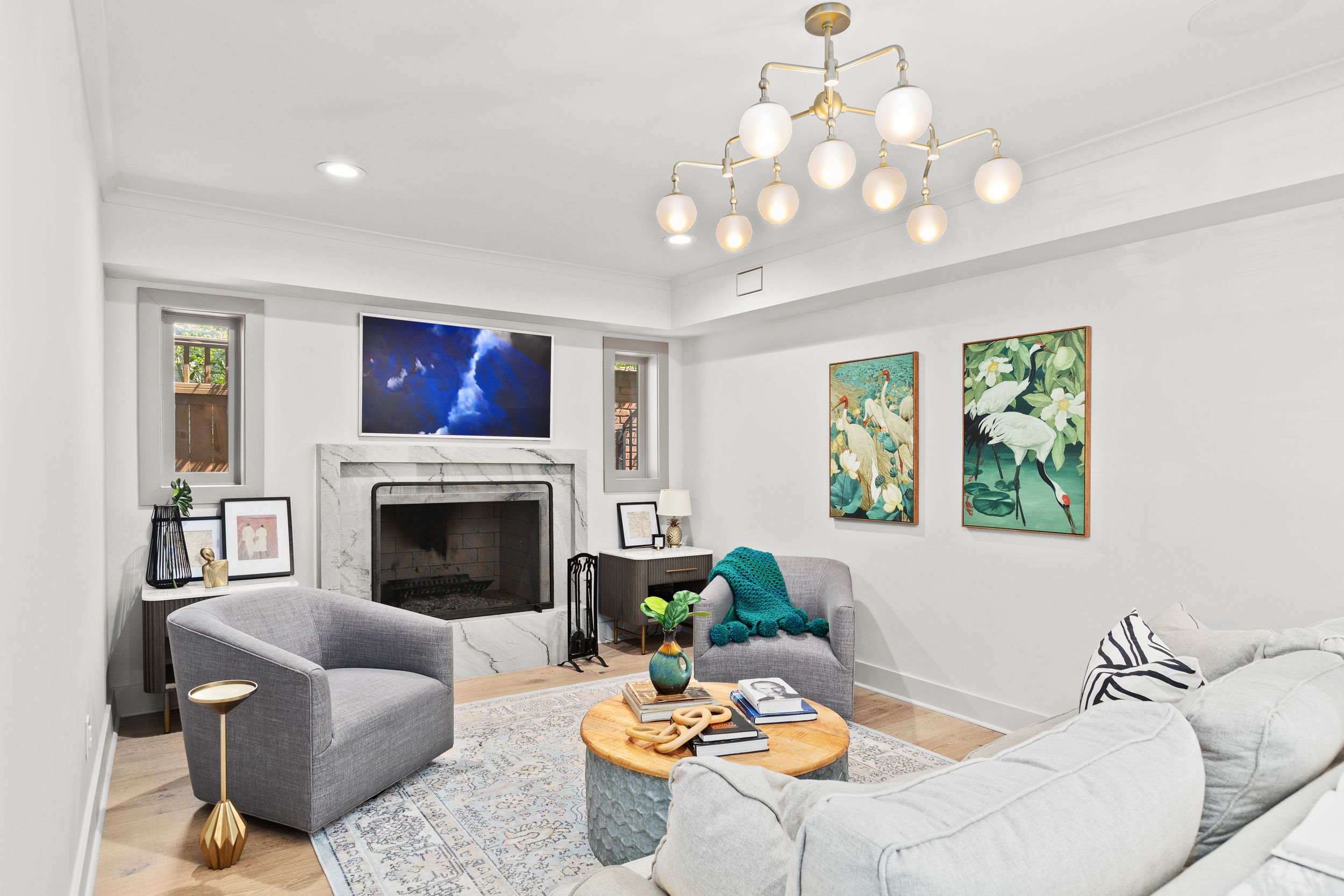 Modern living room features gray sectional, geometric chandelier, fireplace, colorful botanical artwork on light walls. Real estate photo.