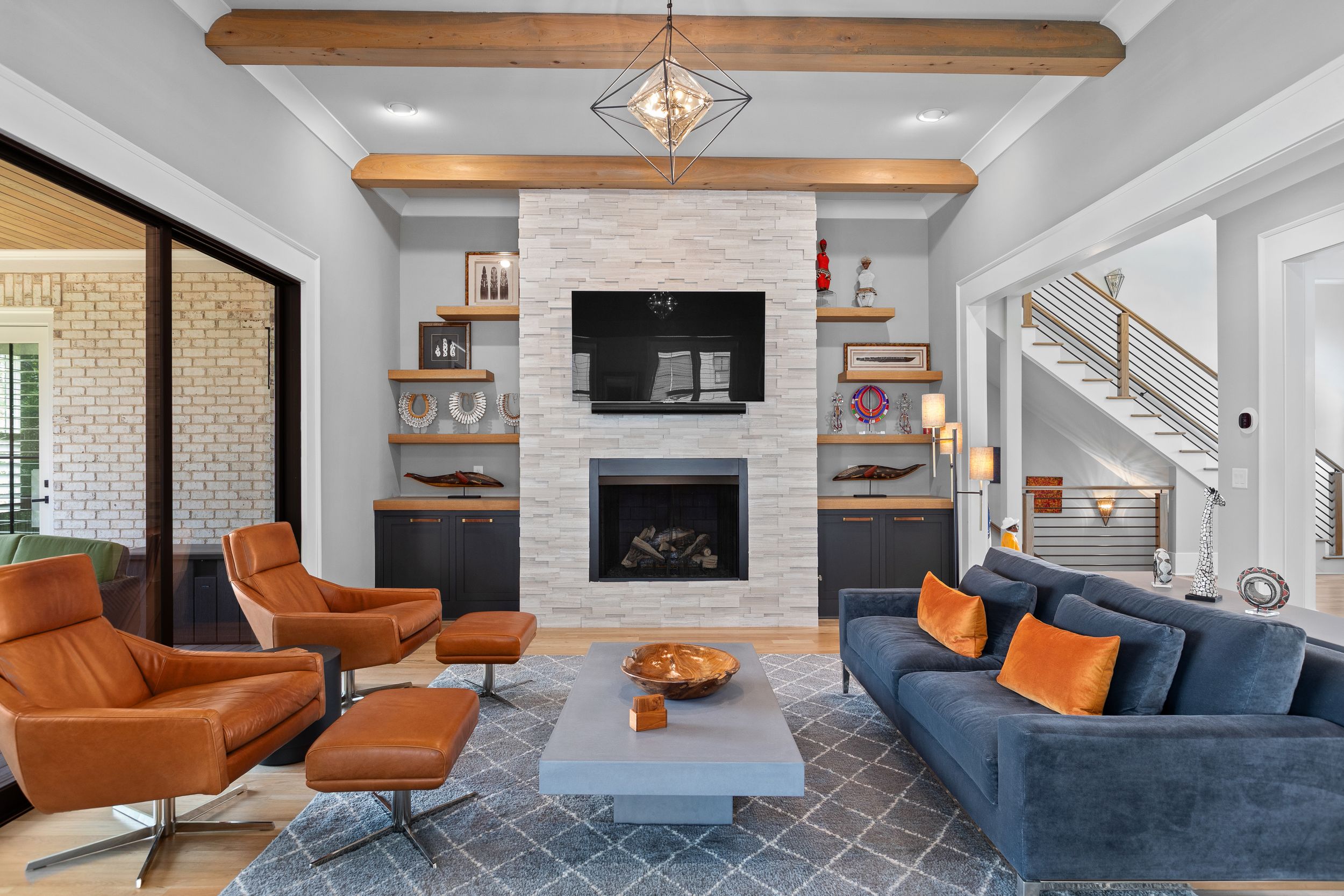 Modern living room interior with navy blue sofa, orange accent chairs, exposed wooden beams and geometric light fixtures. Real estate photo.