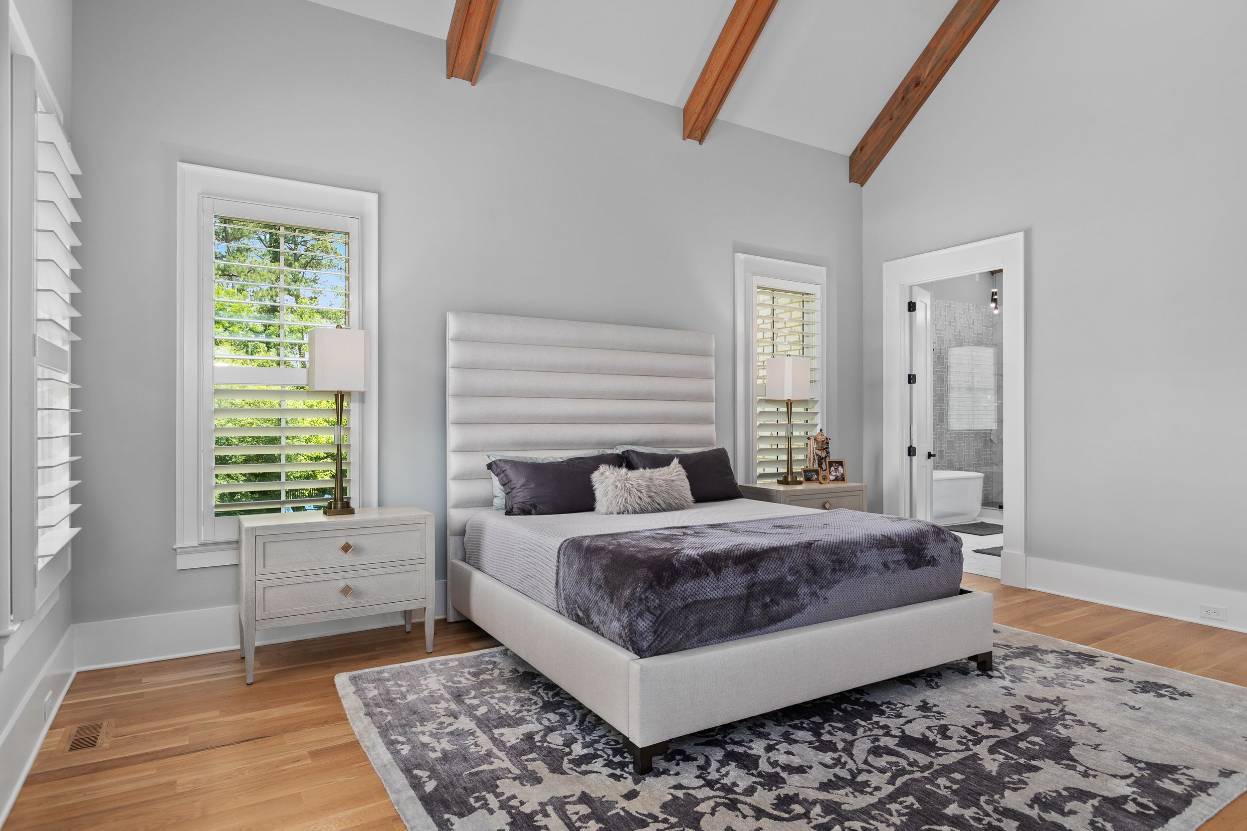 Modern master bedroom with white upholstered bed, gray decor, hardwood floors, and exposed wooden ceiling beams. Real estate photography.