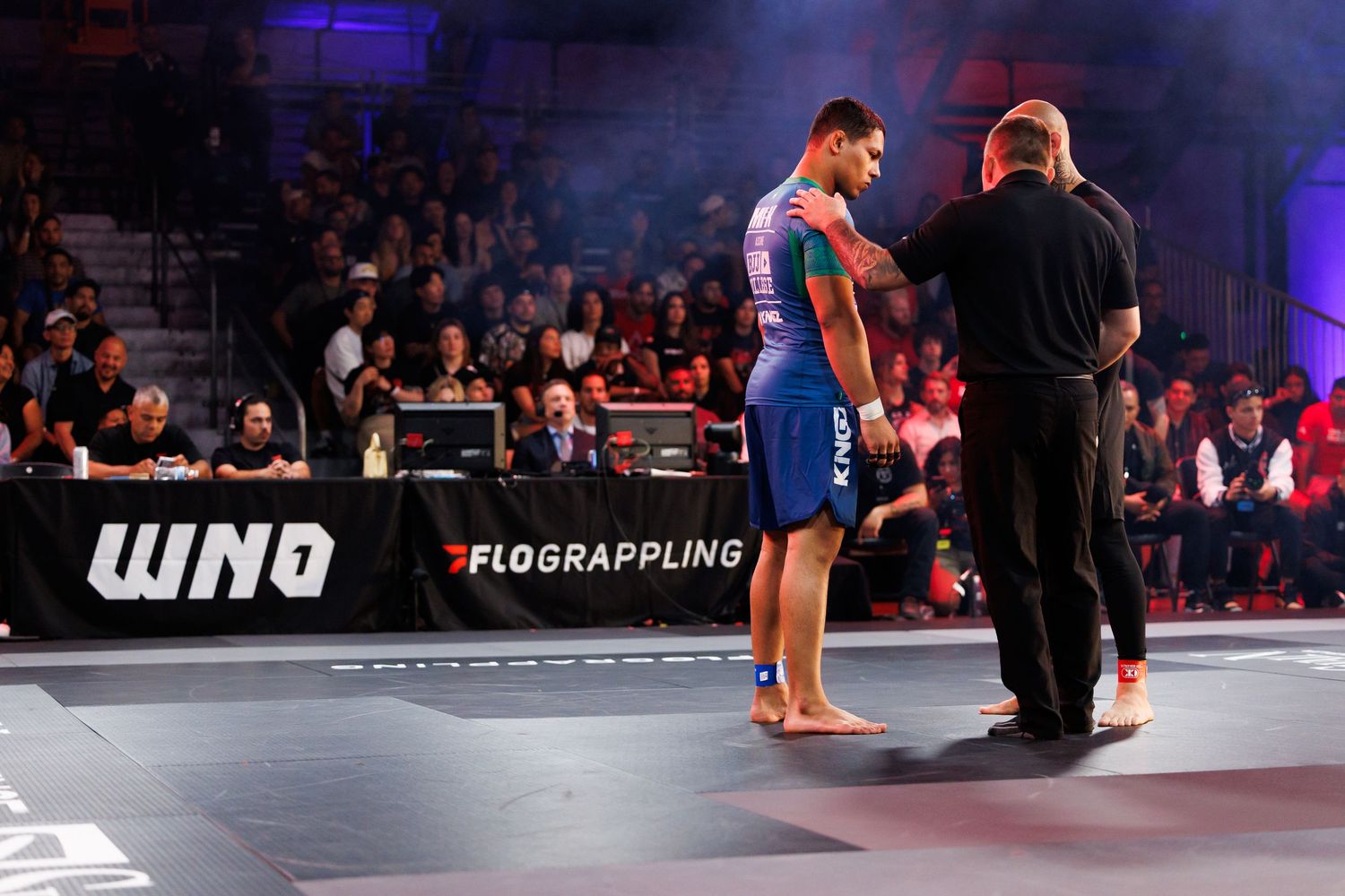 Two martial artists grappling on a blue mat during a competition match.