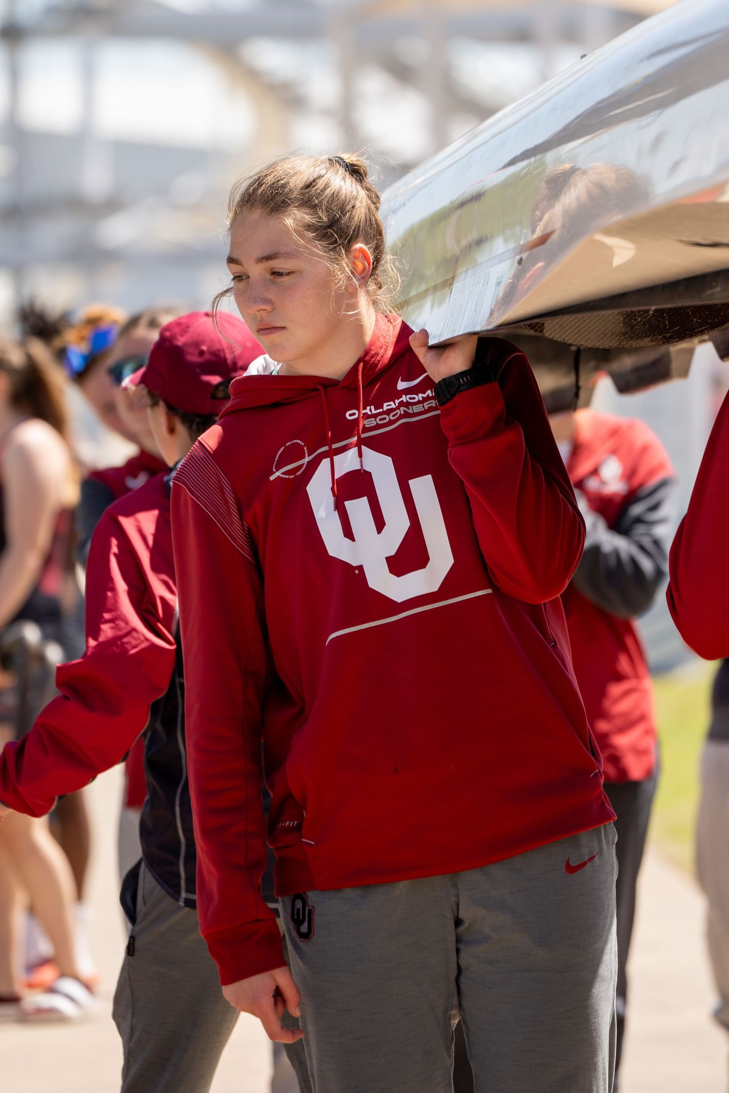 Students in red University of Utah hoodies carry rowing oars on their shoulders while walking in formation outdoors.