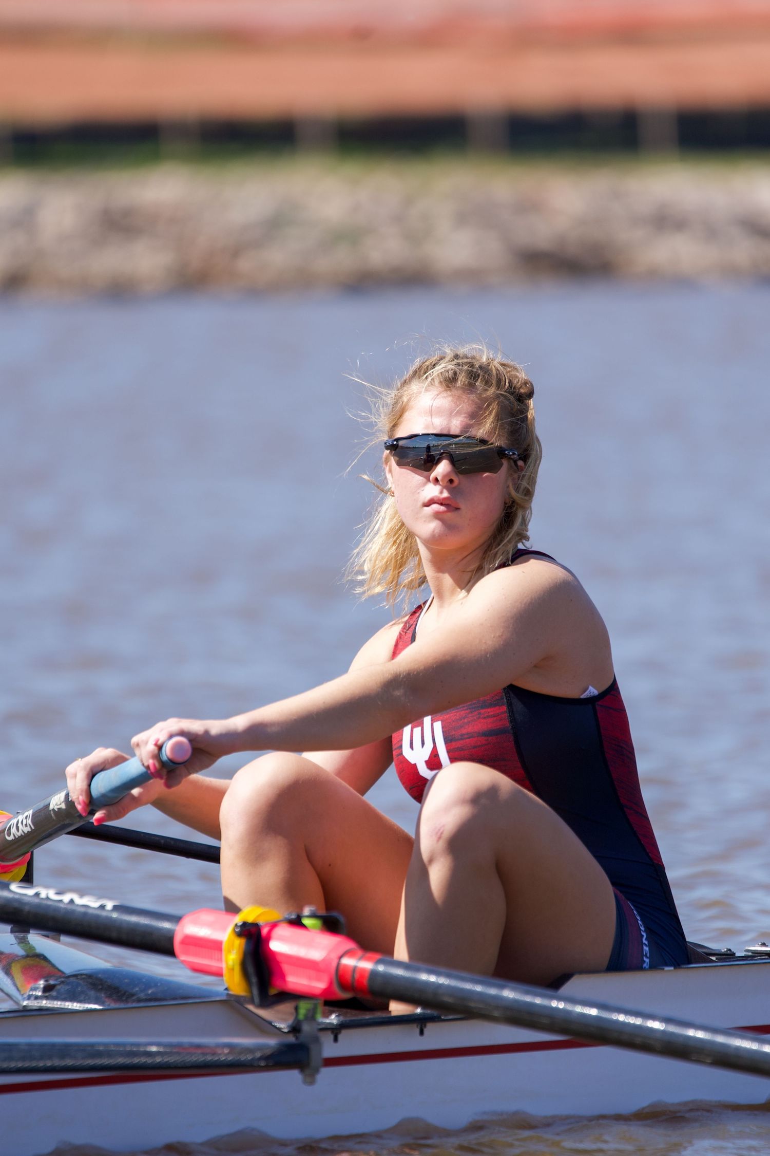 Athlete wearing sunglasses rows single scull boat on calm water wearing navy and red racing uniform.