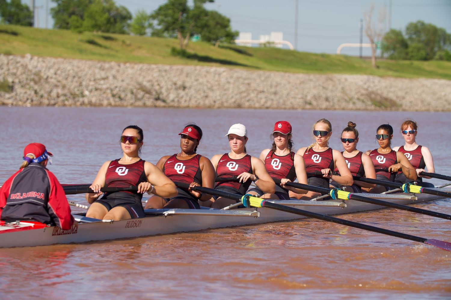 Women's rowing crew team practicing on a river with red accents on their uniforms.