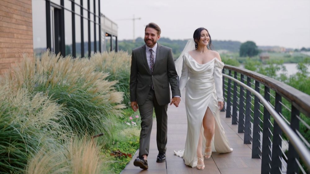 A couple walks together across a wooden walkway surrounded by tall grass at a scenic outdoor venue.
