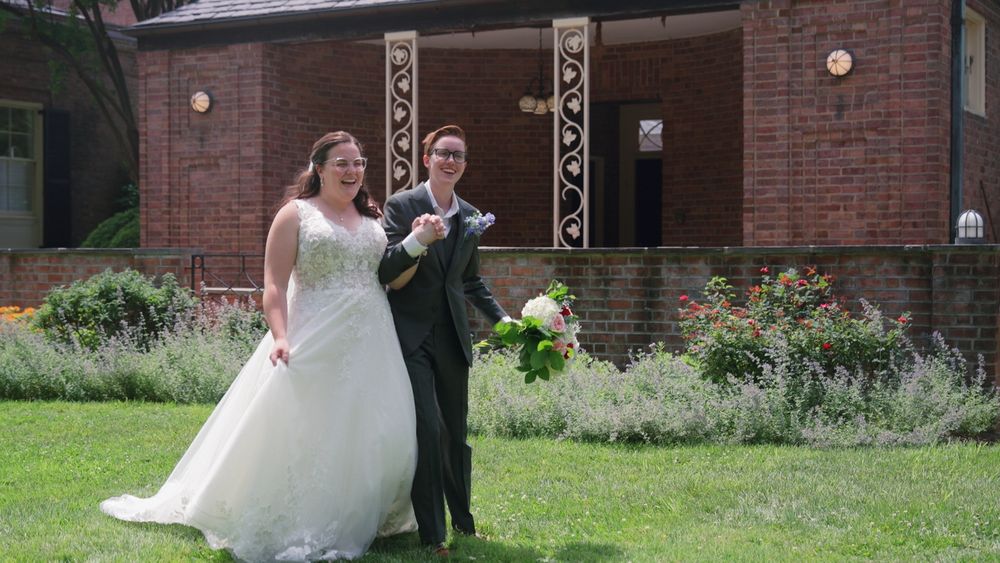 A wedding couple poses together in front of a rustic brick building with landscaped grounds.