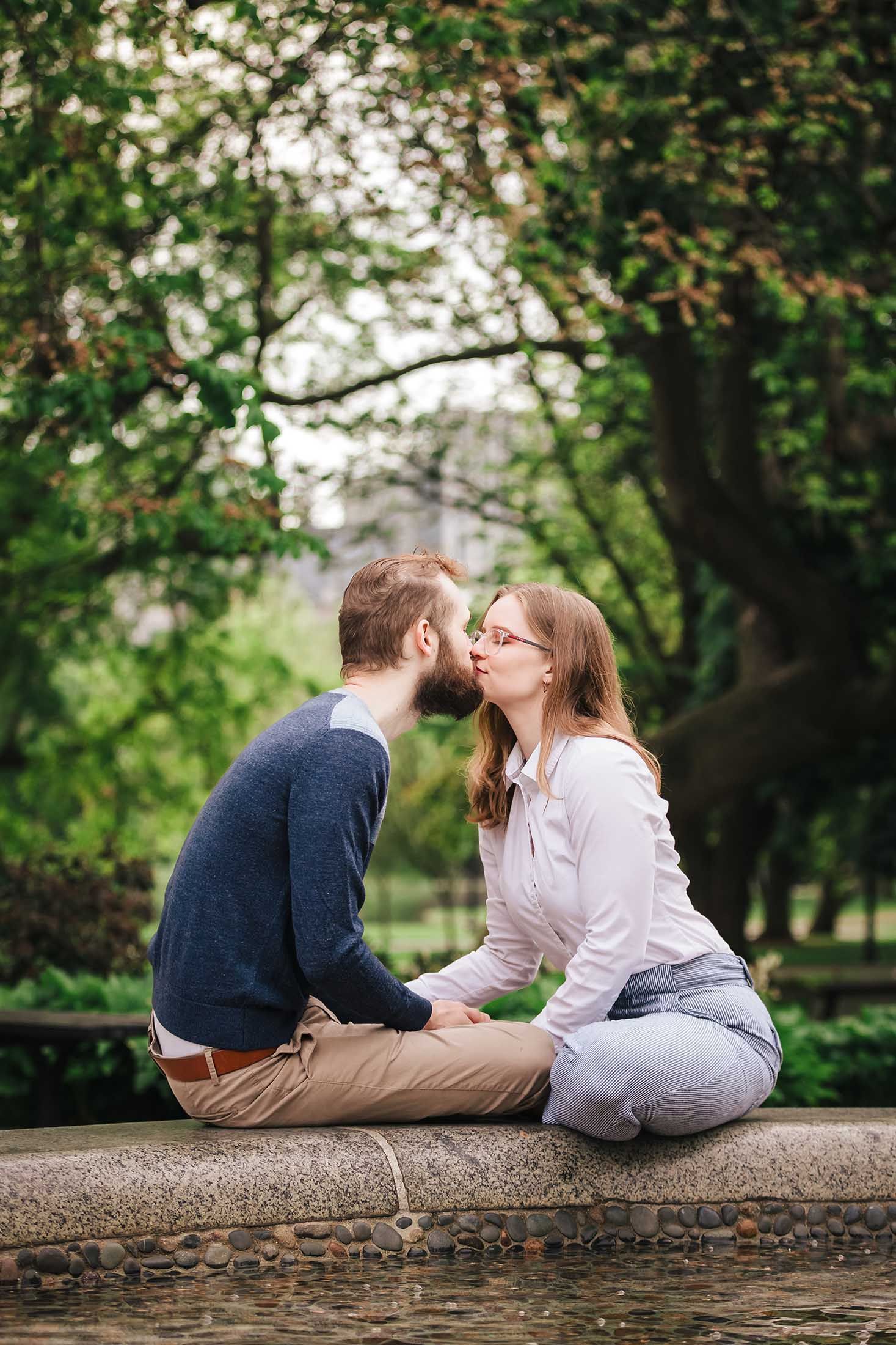 Playful moment of engaged couple sharing a sweet kiss while seated on the edge of the George Robert White Memorial Fountain in Boston Public