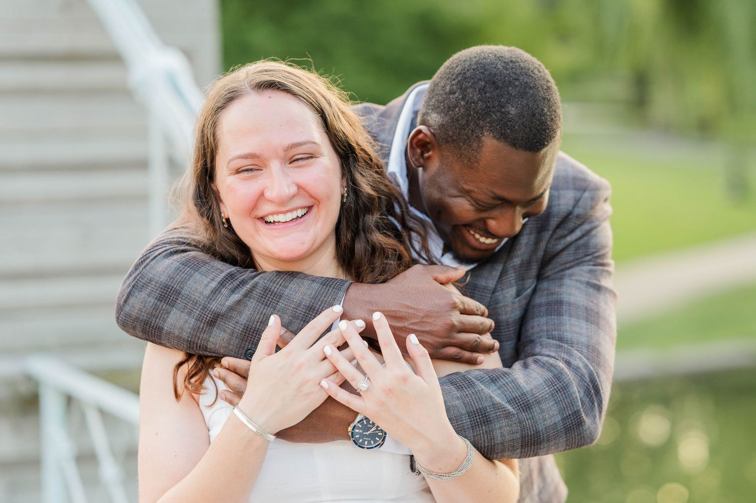 A joyful couple embraces while showing off an engagement ring in an outdoor setting during their engagement photoshoot.