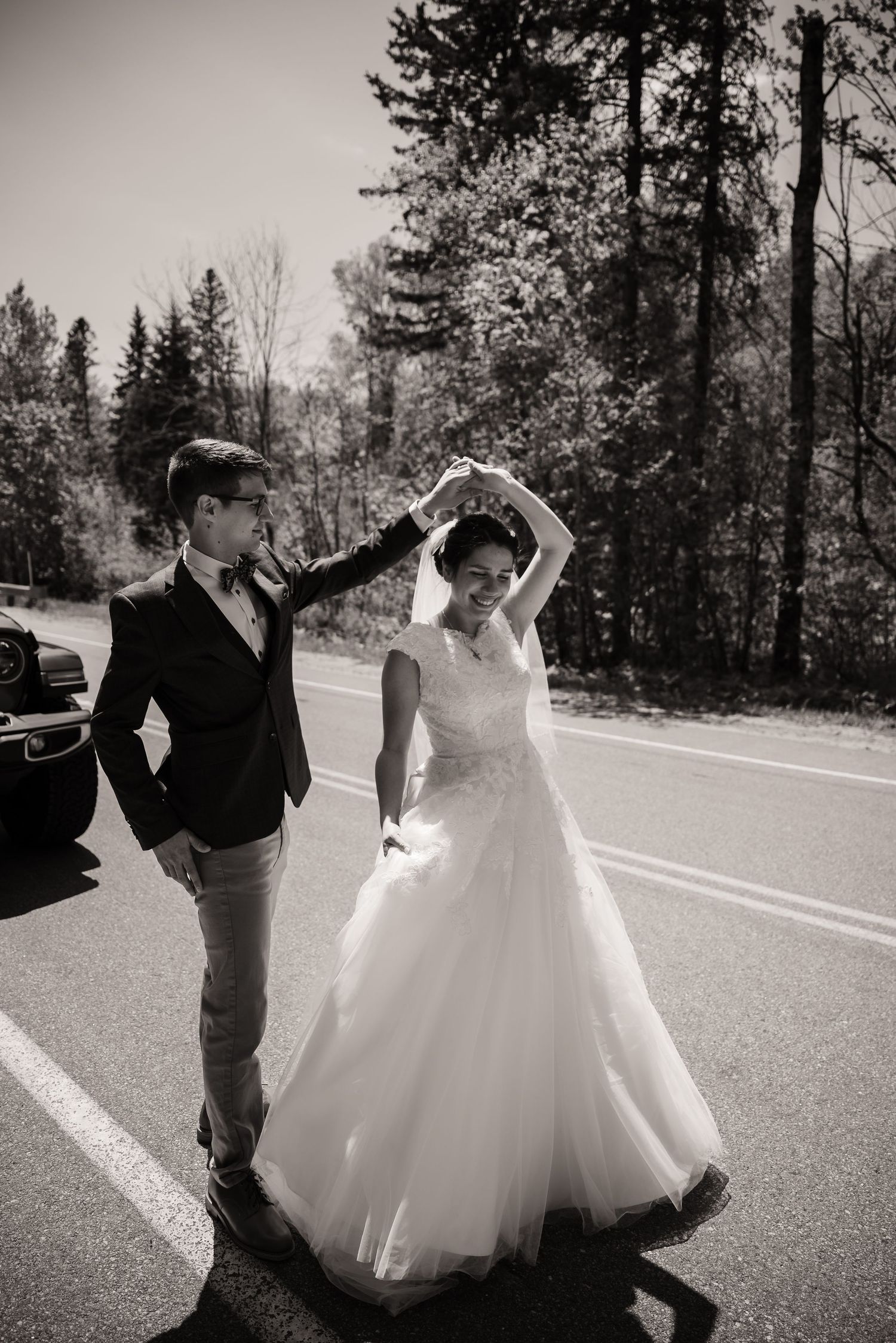 Couple dancing on a mountain road surrounded by evergreen trees in black and white.