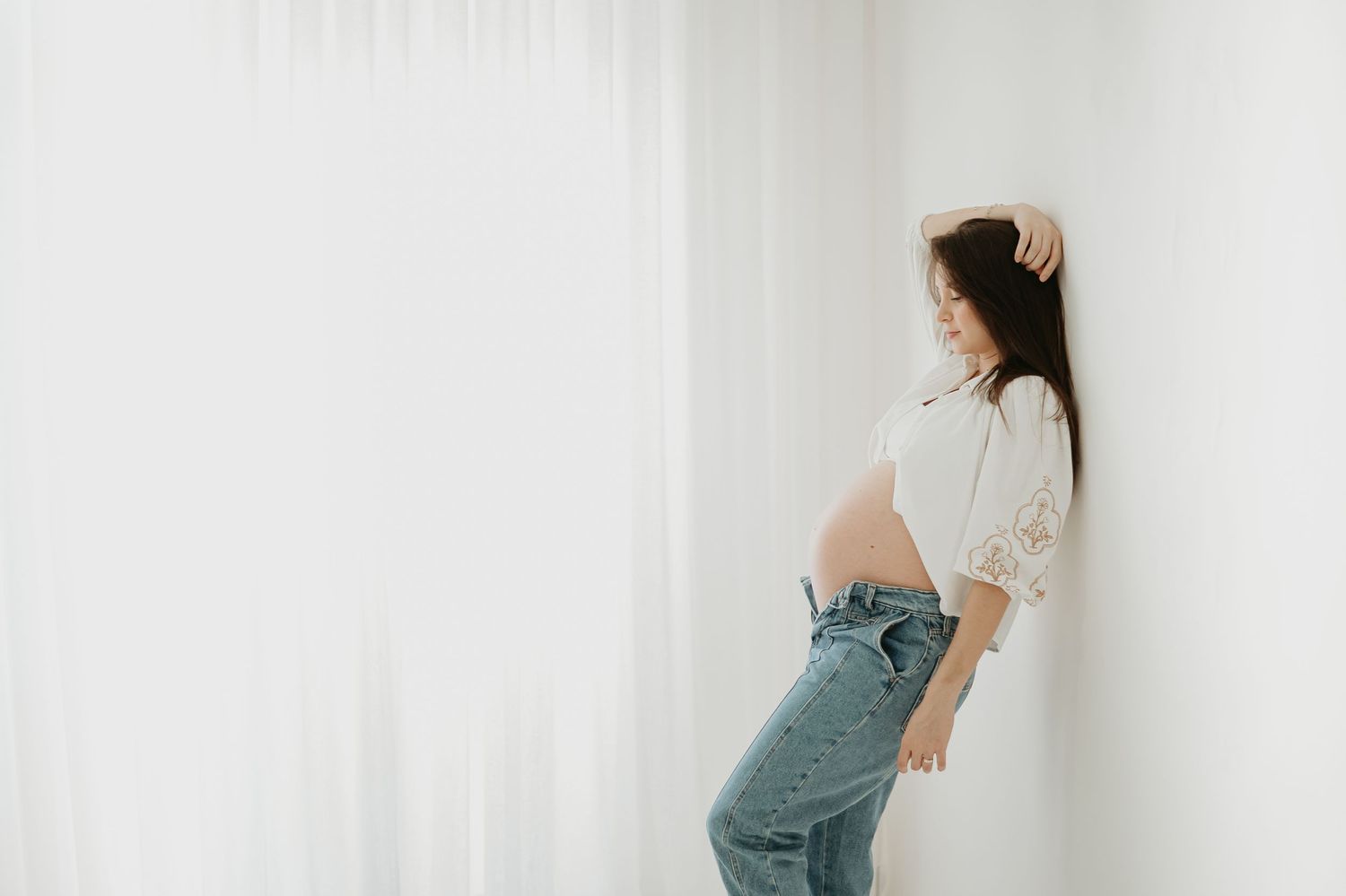 Artistic maternity photo with someone in casual denim against white wall.