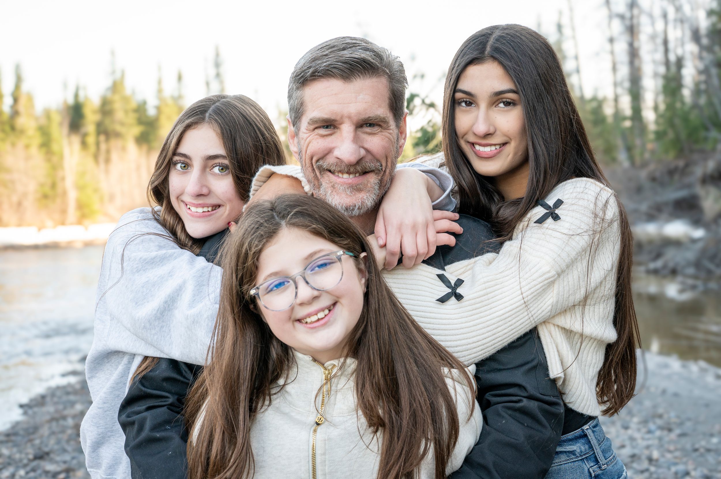 A happy group poses together outdoors on a rocky beach with evergreen trees and sunlight in the background.