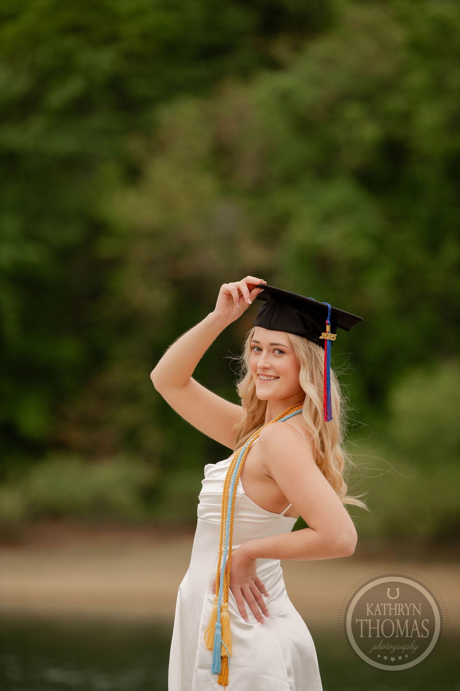 Graduate in white dress adjusts cap against blurred green background during outdoor photoshoot.