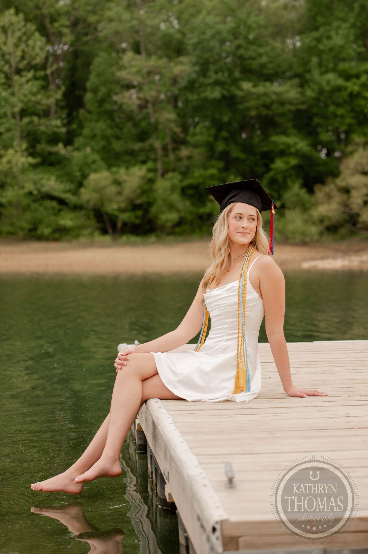 Graduate in white dress and cap sits on wooden dock by water wearing gold and blue honor cords.