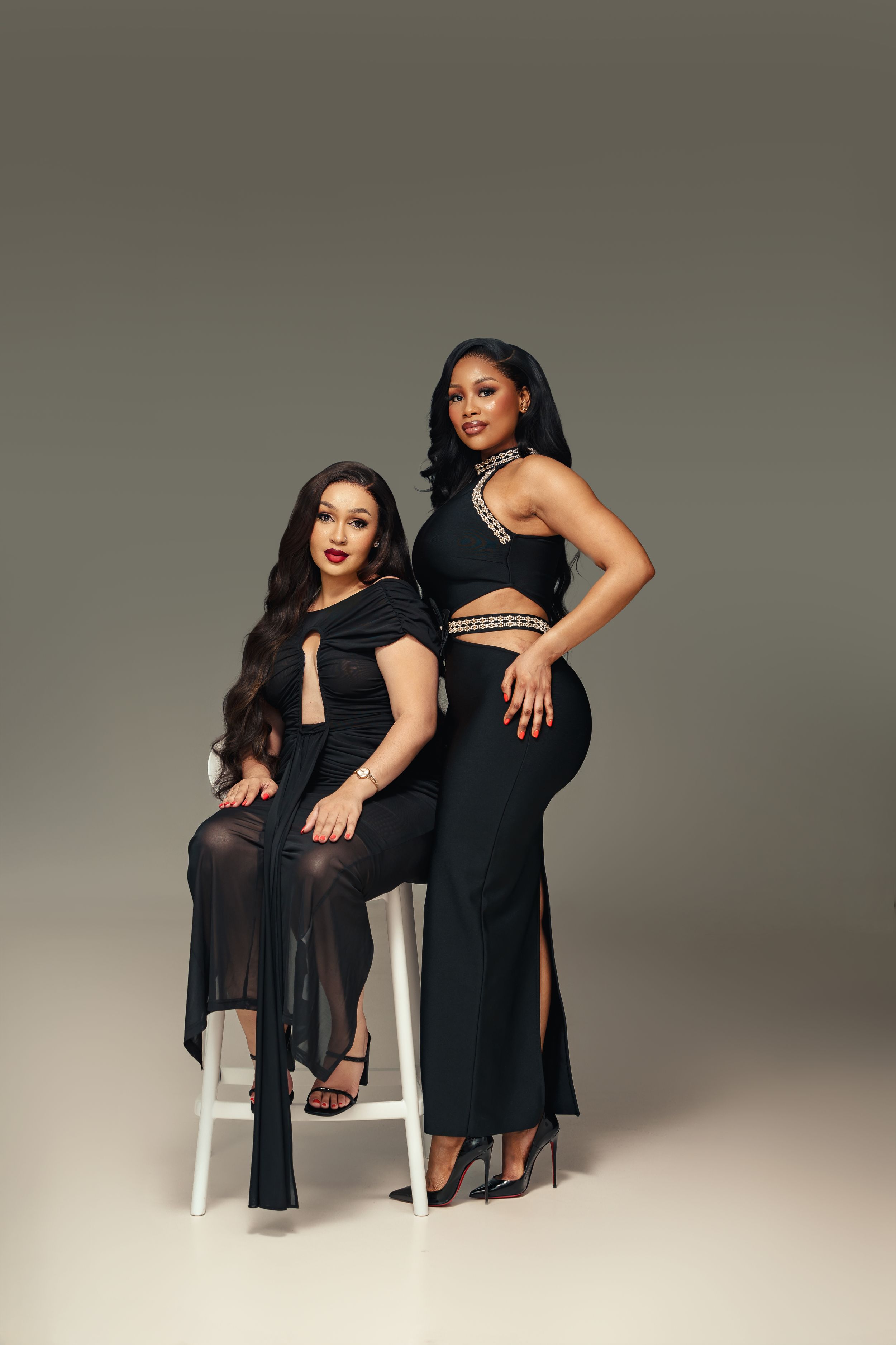 Two women in elegant black evening gowns posing together against a neutral backdrop, one seated on a white stool.