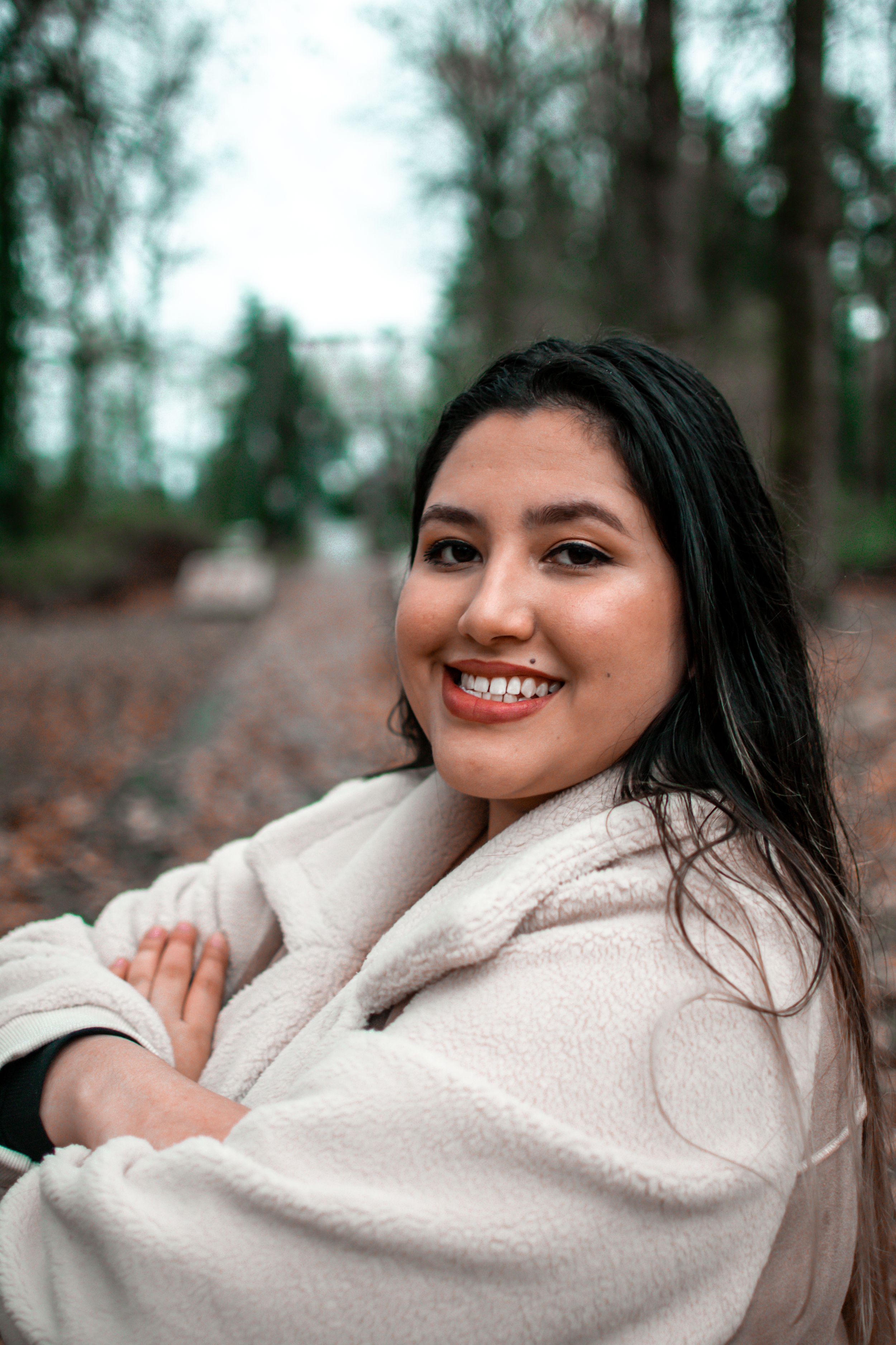 Someone wearing a cozy cream-colored winter coat sits outdoors on fallen autumn leaves with a warm smile.