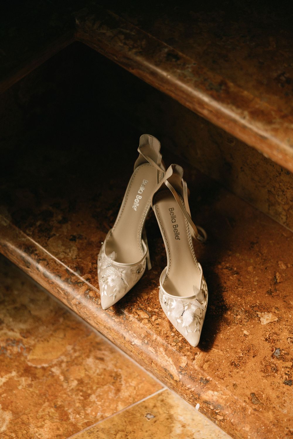 Lace pointed-toe bridal heels with bow detail placed on rustic stone steps in warm light.