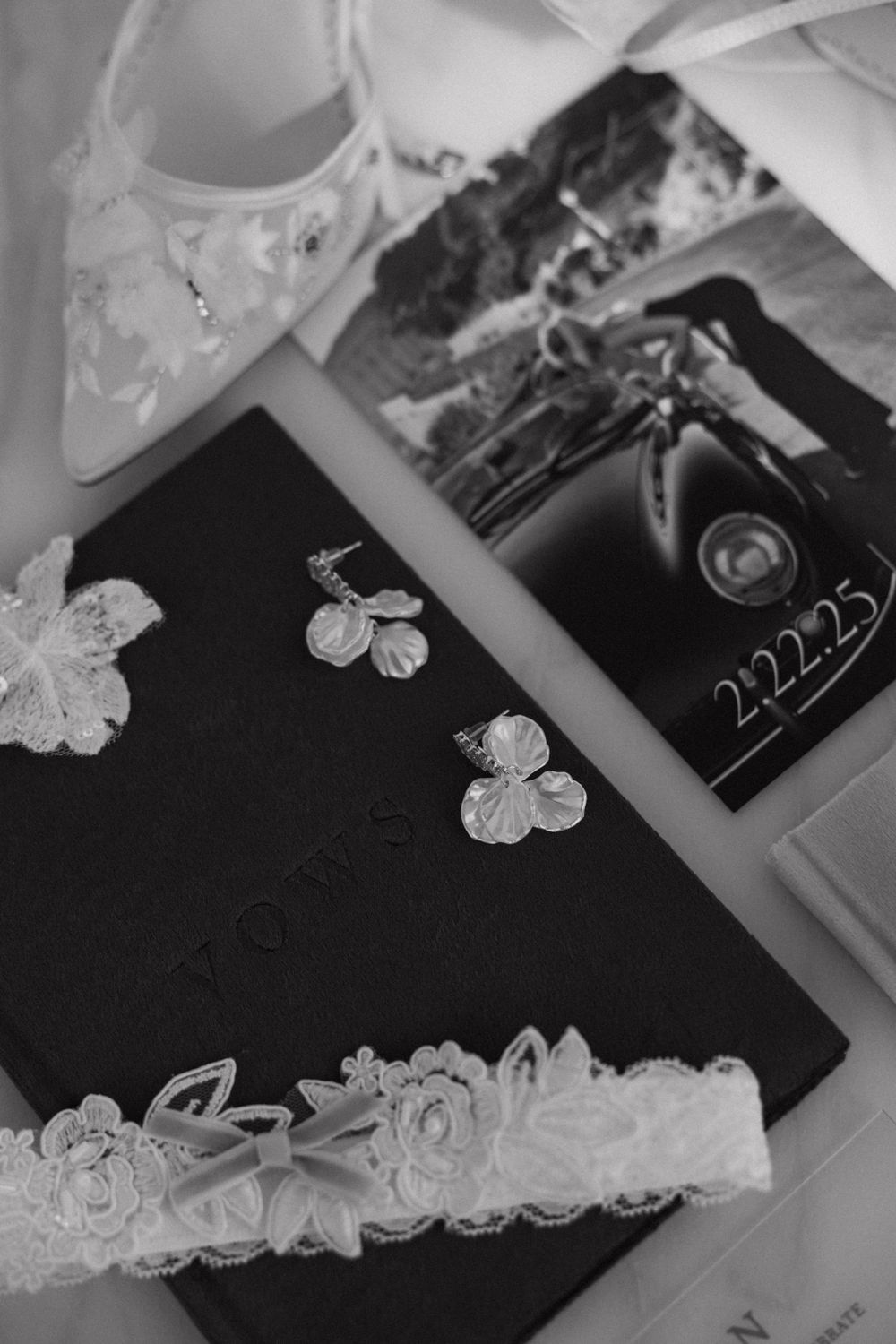 Black and white flat lay of bridal accessories including lace garter, earrings, and perfume box.