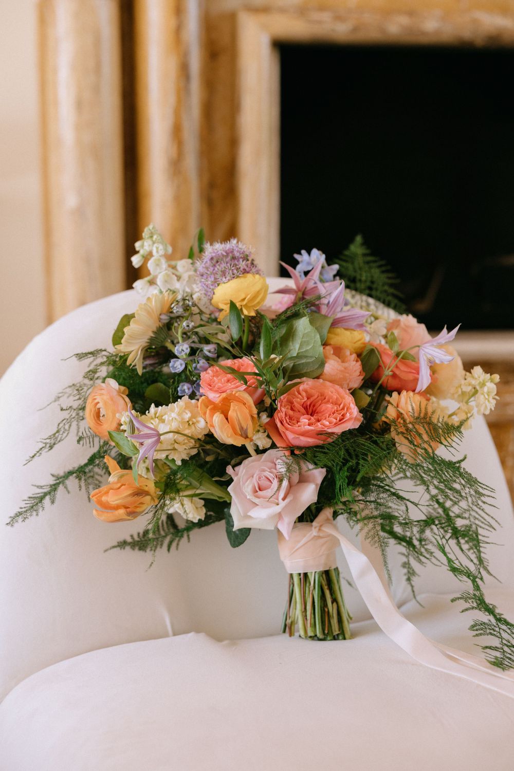 Colorful bridal bouquet with peach roses, orange ranunculus, and purple flowers on white chair.