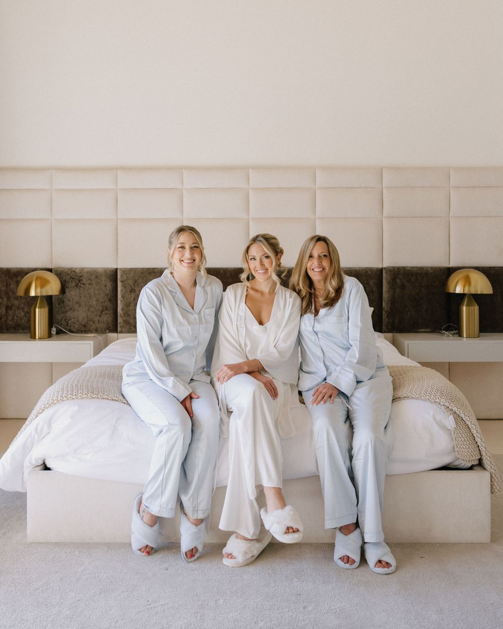 Bride and two bridesmaids in matching pajamas smiling on white bed with gold bedside lamps.